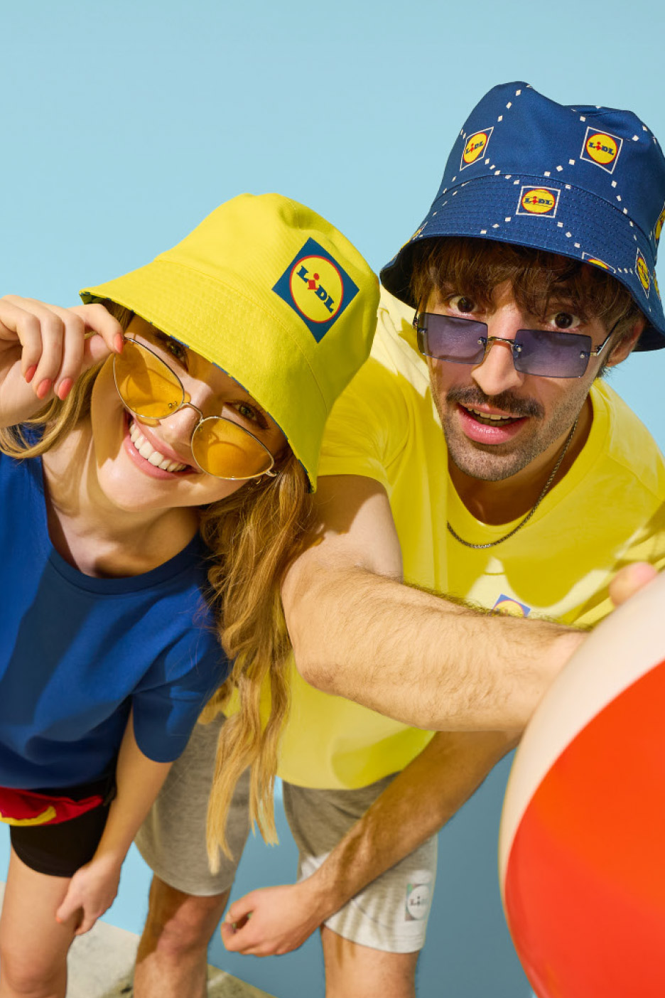 Two people in bucket hats and sunglasses, one yellow, one blue, with a beach ball.