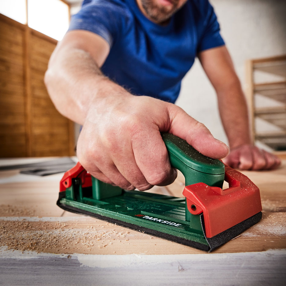 Man sanding a wooden surface with a hand sander, with sawdust around.