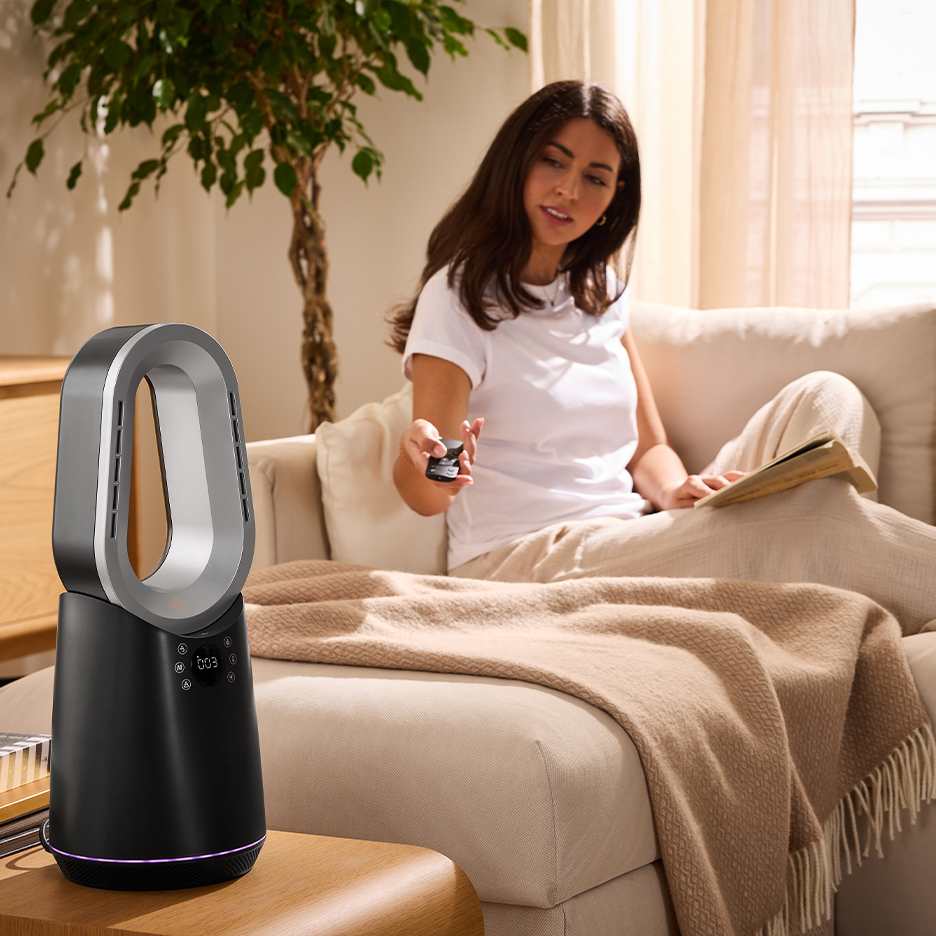 A woman on a sofa uses a remote to control a black and silver bladeless fan.