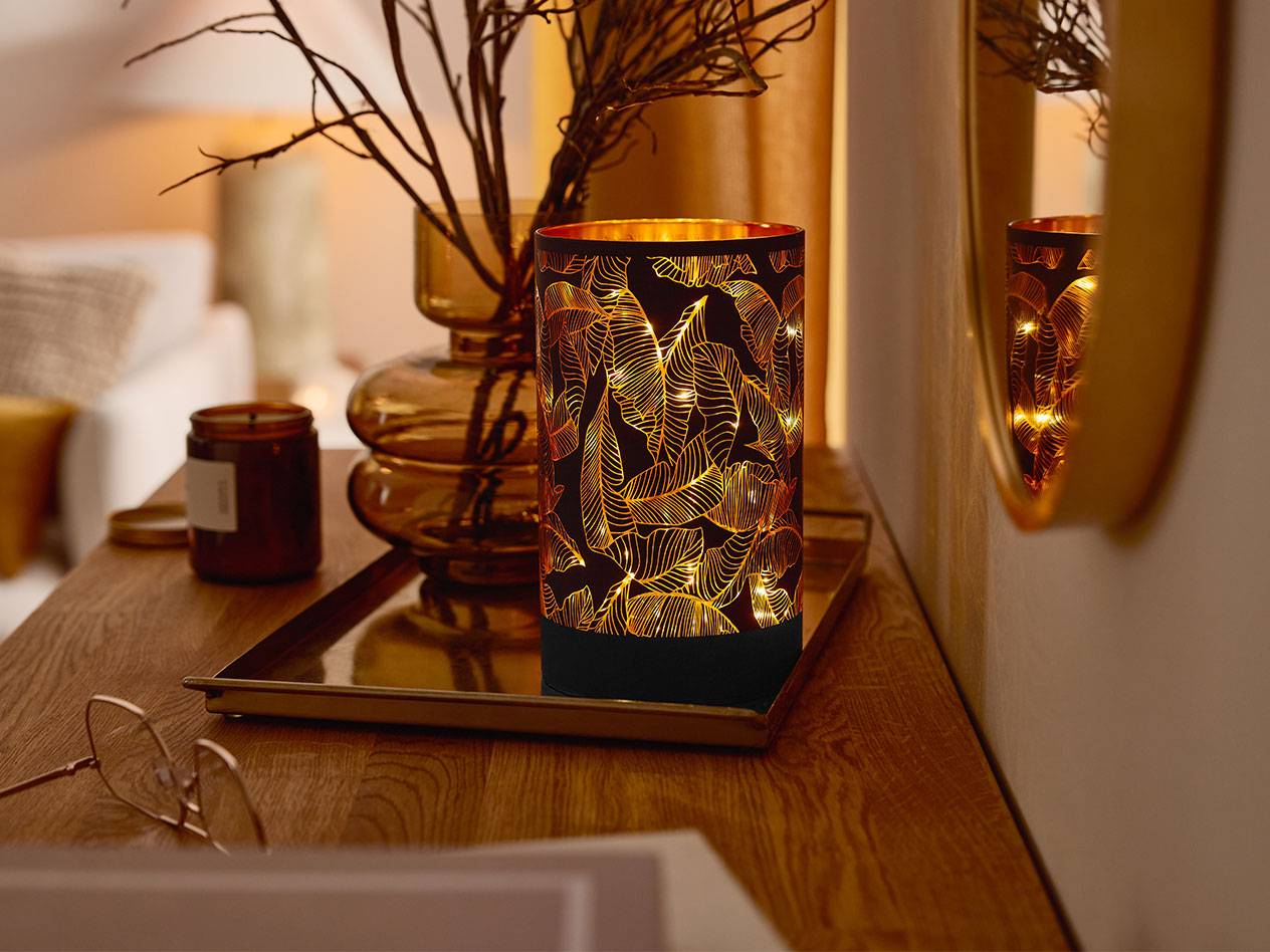 A decorative LED lantern with a leaf pattern, a vase with branches, and a candle on a wooden table.
