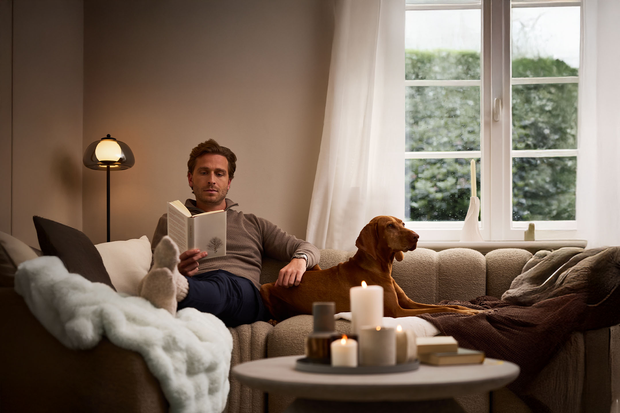 Man reading book with dog on sofa, cozy lighting and scented candles.