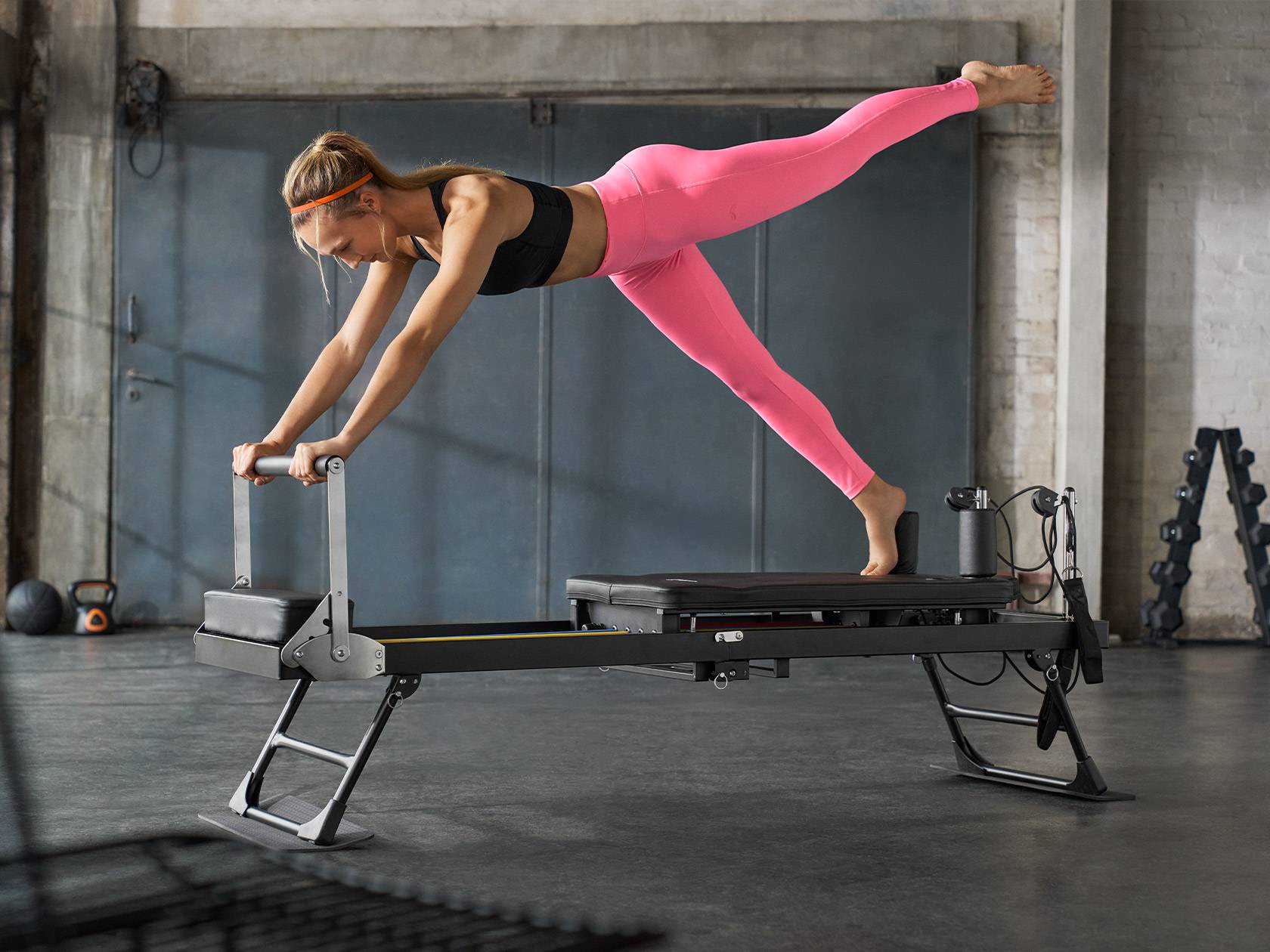 Woman in pink leggings exercising on a Pilates reformer machine in a gym.
