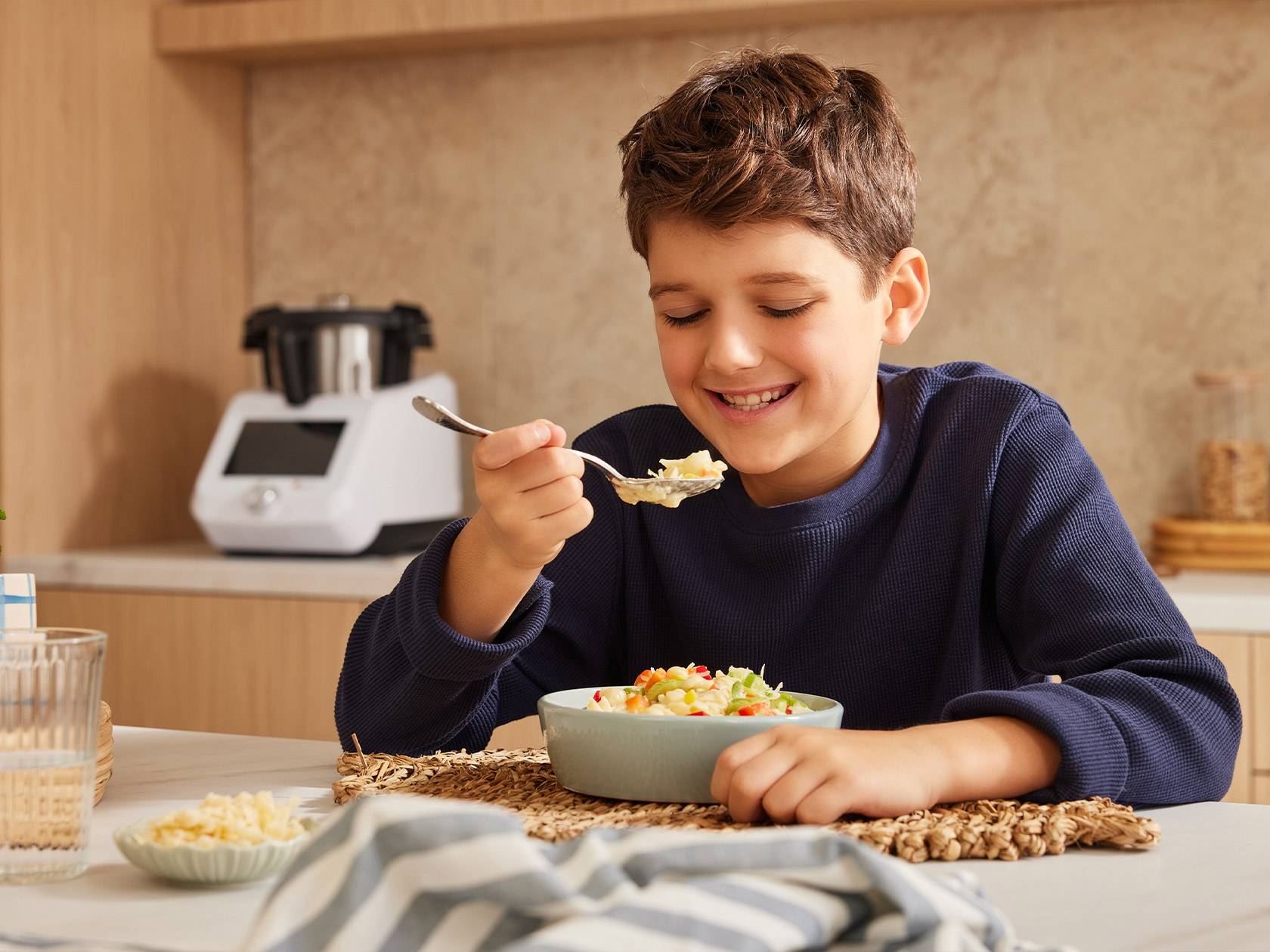 A smiling boy eating from a bowl with a kitchen appliance in the background.
