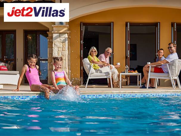 Two girls splashing in a pool while adults relax on a patio, with a villa in the background.