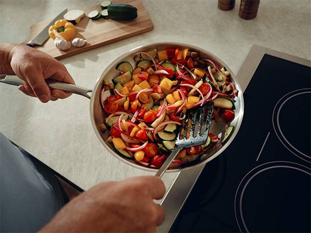 Frying pan with chopped vegetables like zucchini, bell peppers, onions, and tomatoes on an induction hob.