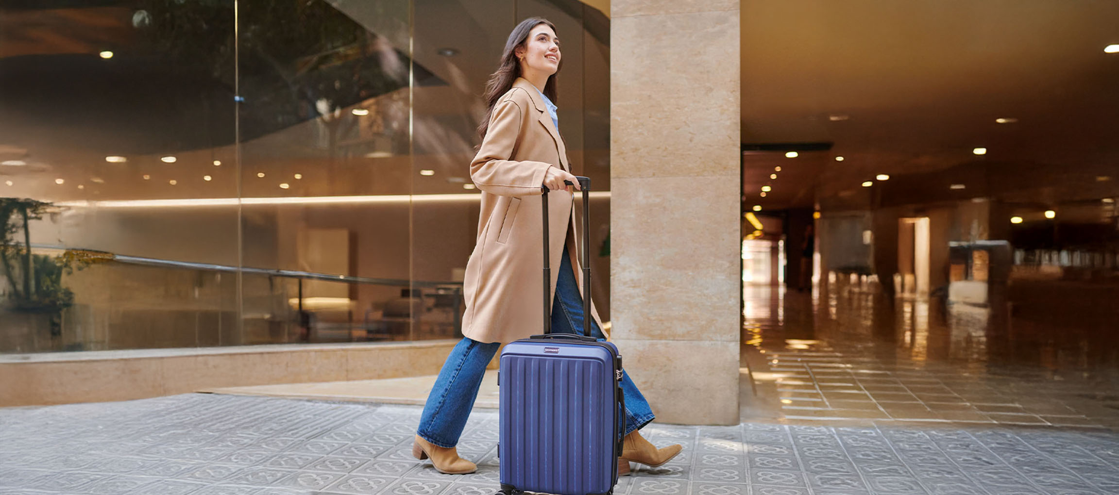 Woman in a trench coat pulling a blue suitcase on patterned pavement.