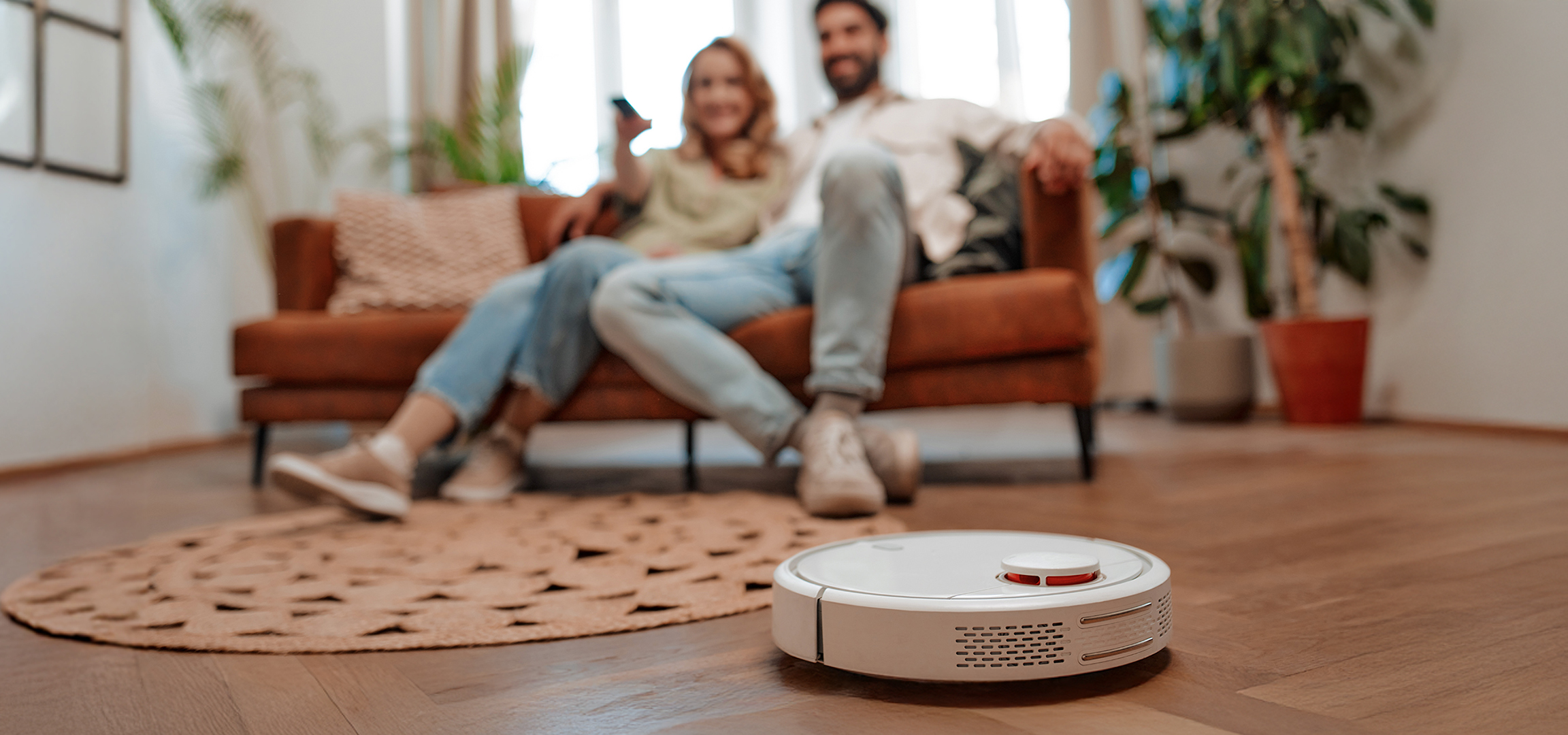 A golden retriever lies on a rug while a robot vacuum cleans the wooden floor.