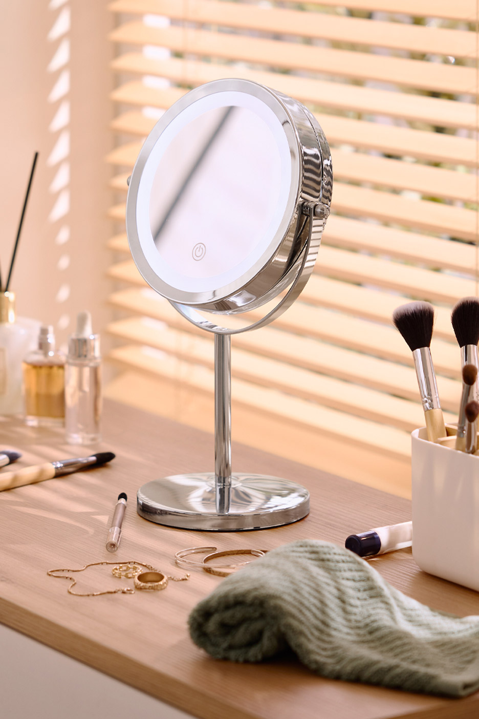 Lighted makeup mirror on a wooden vanity with cosmetics, jewelry, and a green towel.
