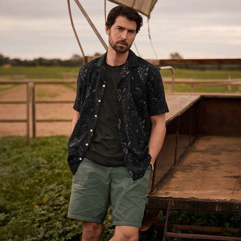 Man in black patterned shirt, t-shirt, and green shorts, standing by a trailer.