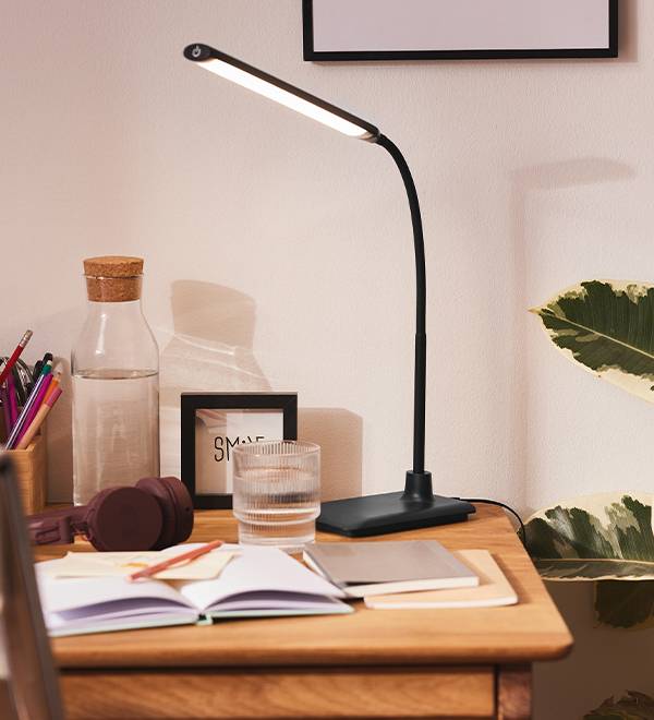 A black desk lamp illuminates a wooden desk with a notebook, water bottle, and headphones.