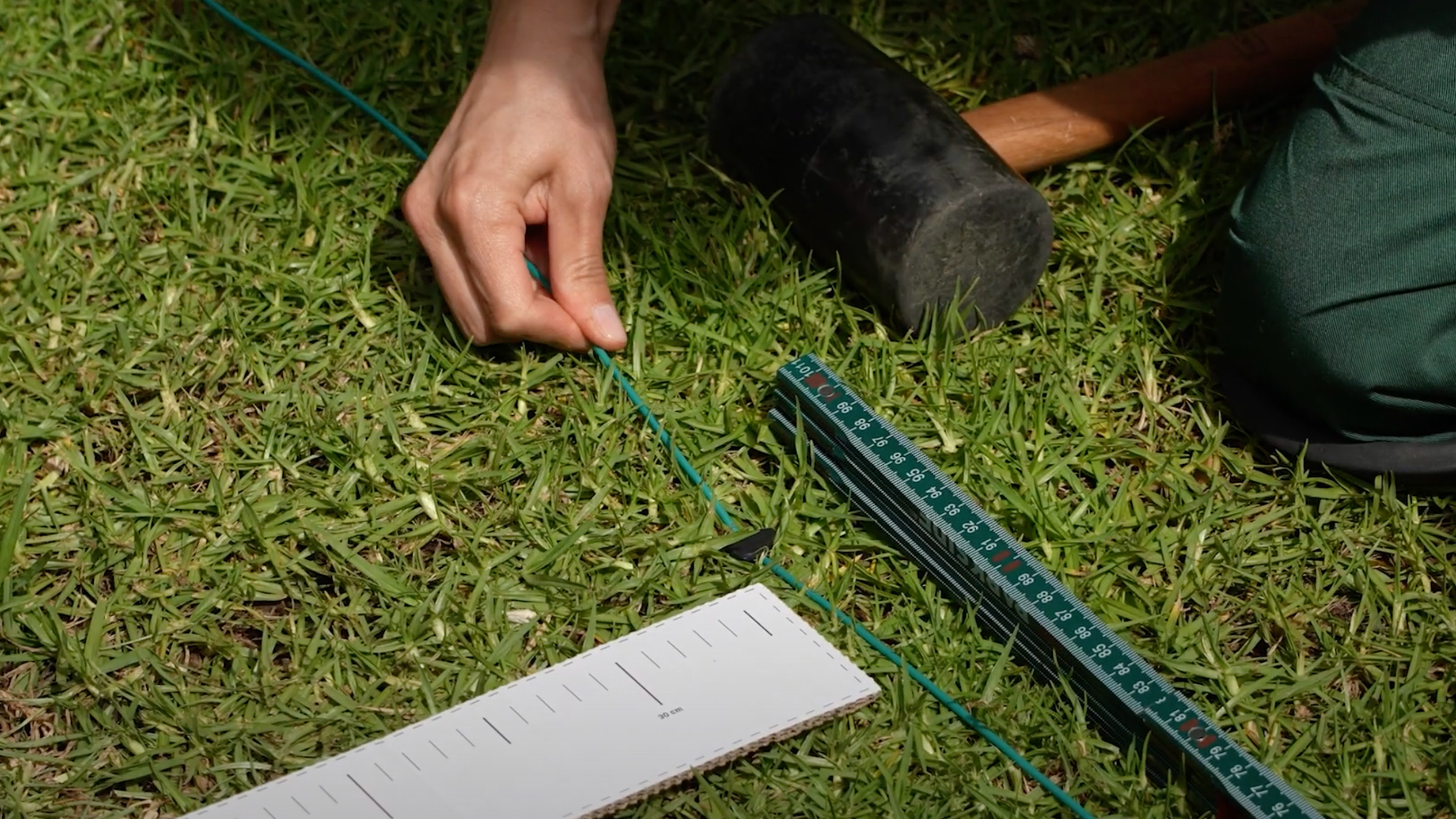 A person uses a mallet and measuring tools to install boundary wire for a robotic lawnmower.