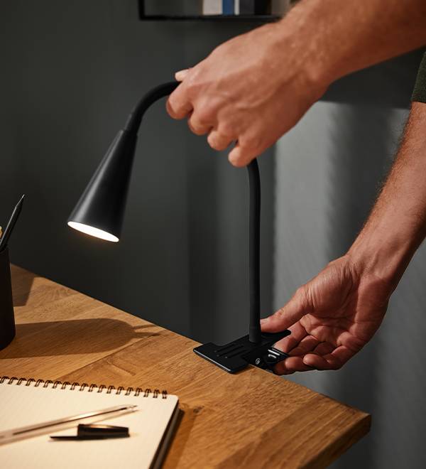 A person adjusting a black clip-on desk lamp on a wooden desk.