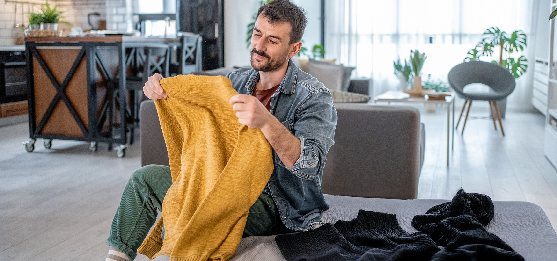 Man holding a yellow knitted sweater, with other sweaters on a sofa in a living room.