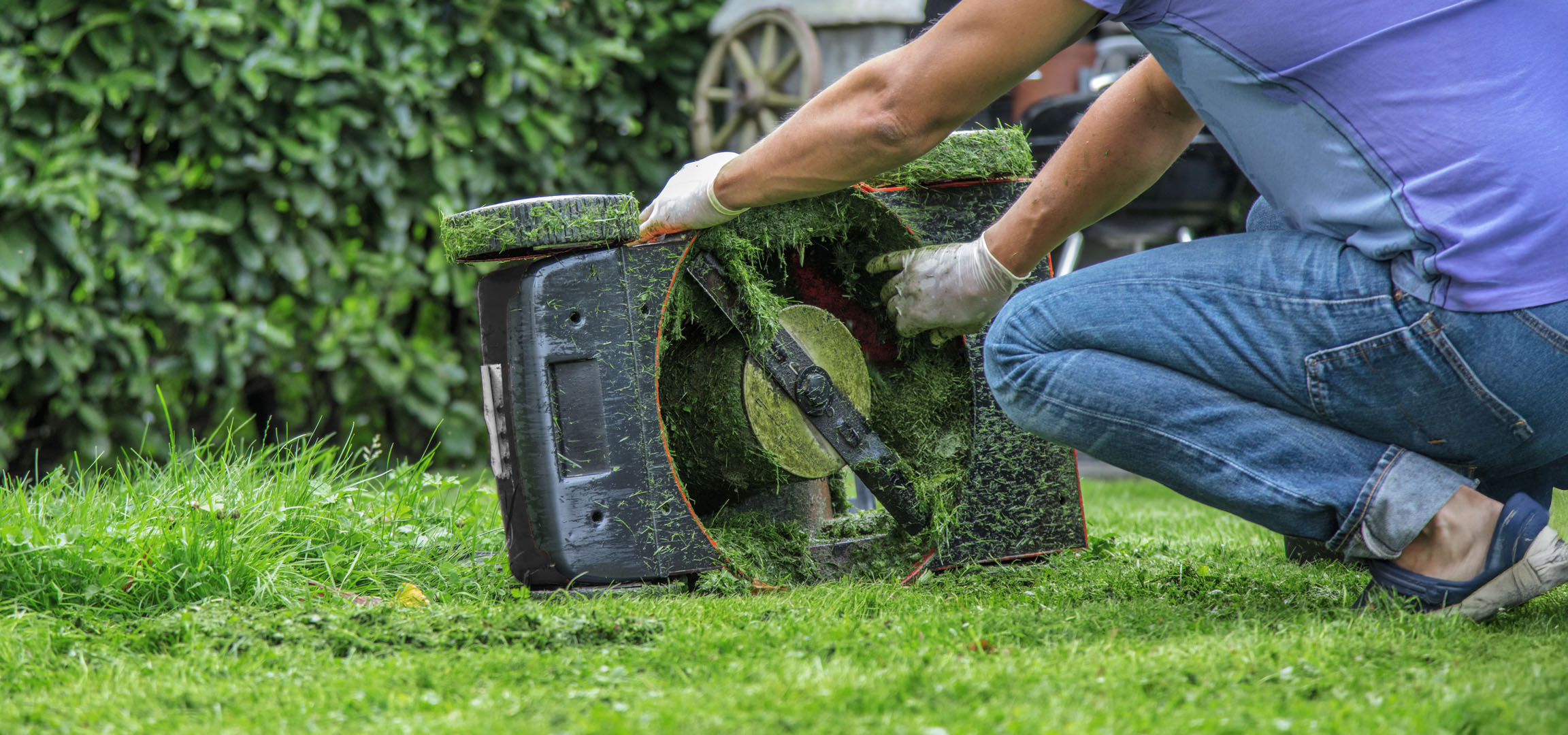 Person cleaning a lawnmower covered in fresh grass clippings in a garden.