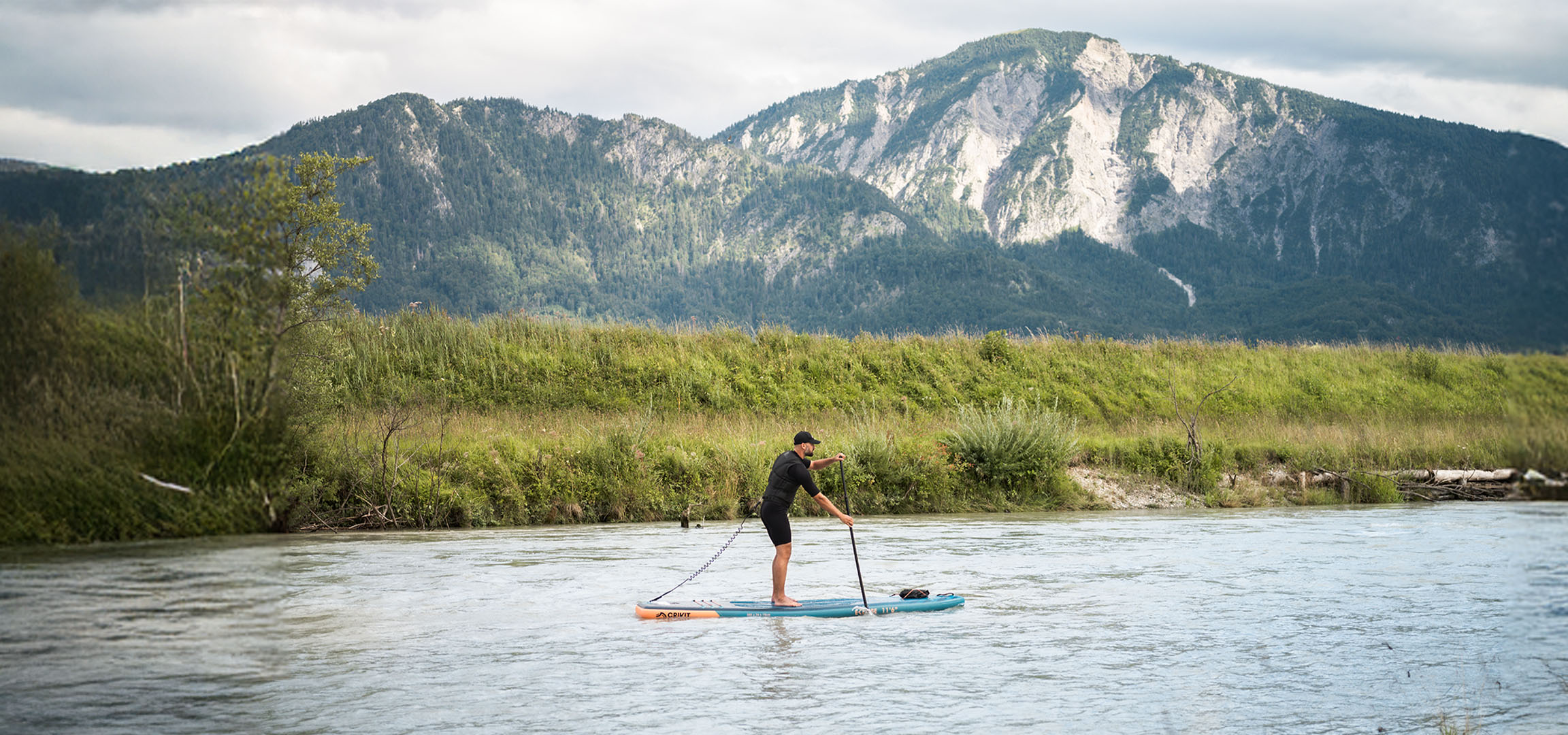 Man paddleboarding on a river with mountains and lush vegetation in the background.