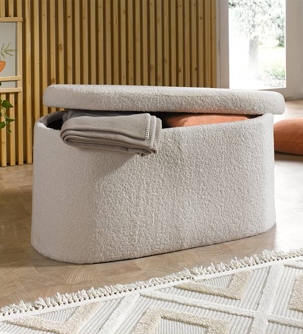 A cream-colored storage ottoman with a blanket and pillows inside, on a wooden floor.