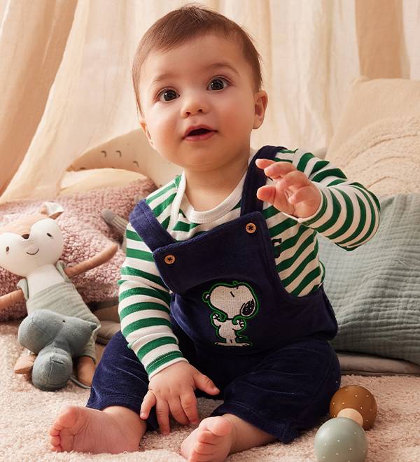 Baby in Snoopy overalls and striped shirt, sitting on a rug with toys.