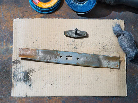 Rusty lawnmower blade on cardboard with abrasive discs and steel wool.