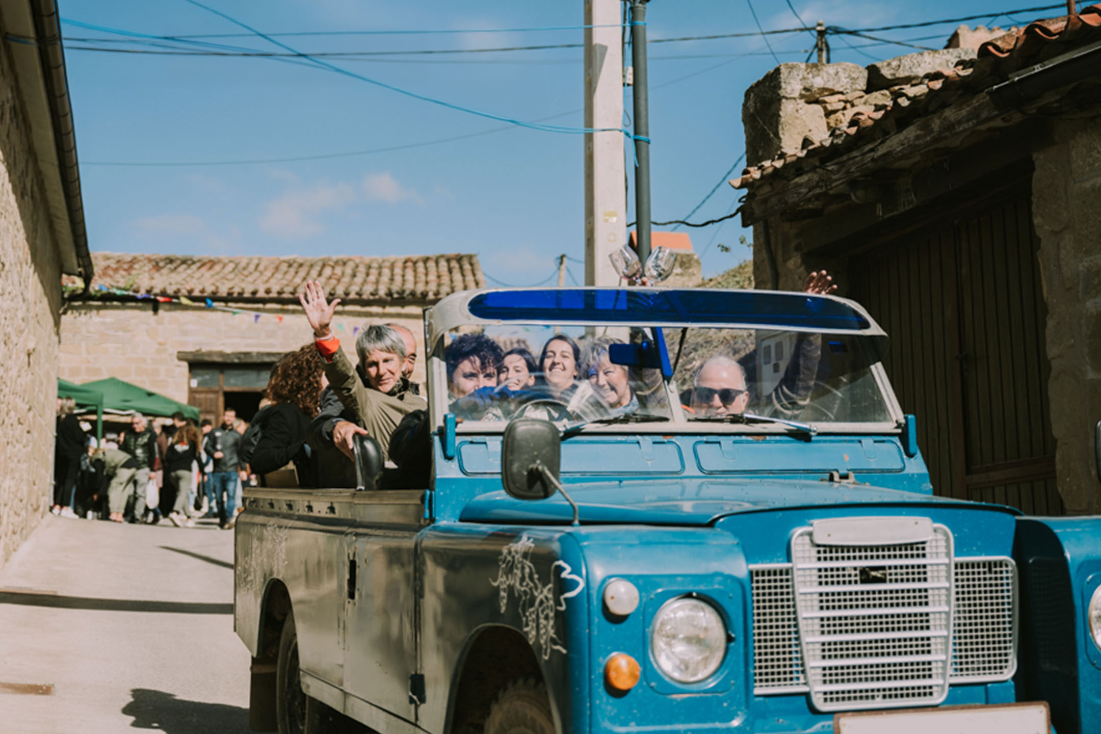People in a blue off-road vehicle on a sunny day, with buildings and a crowd in the background.