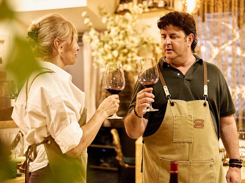 Two people tasting red wine, wearing aprons.