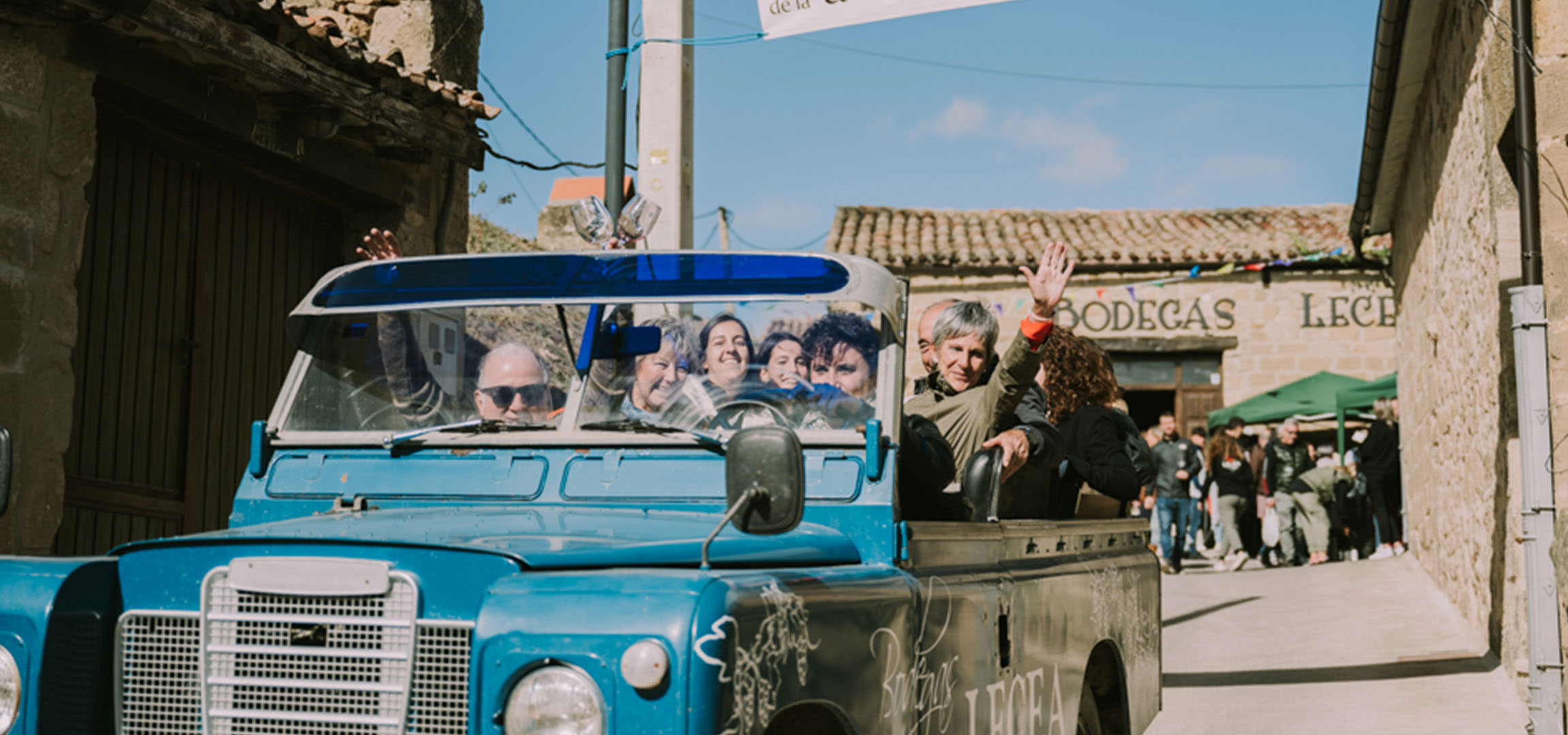 People in a blue off-road vehicle passing by a winery with the inscription 'BODEGAS LECE'