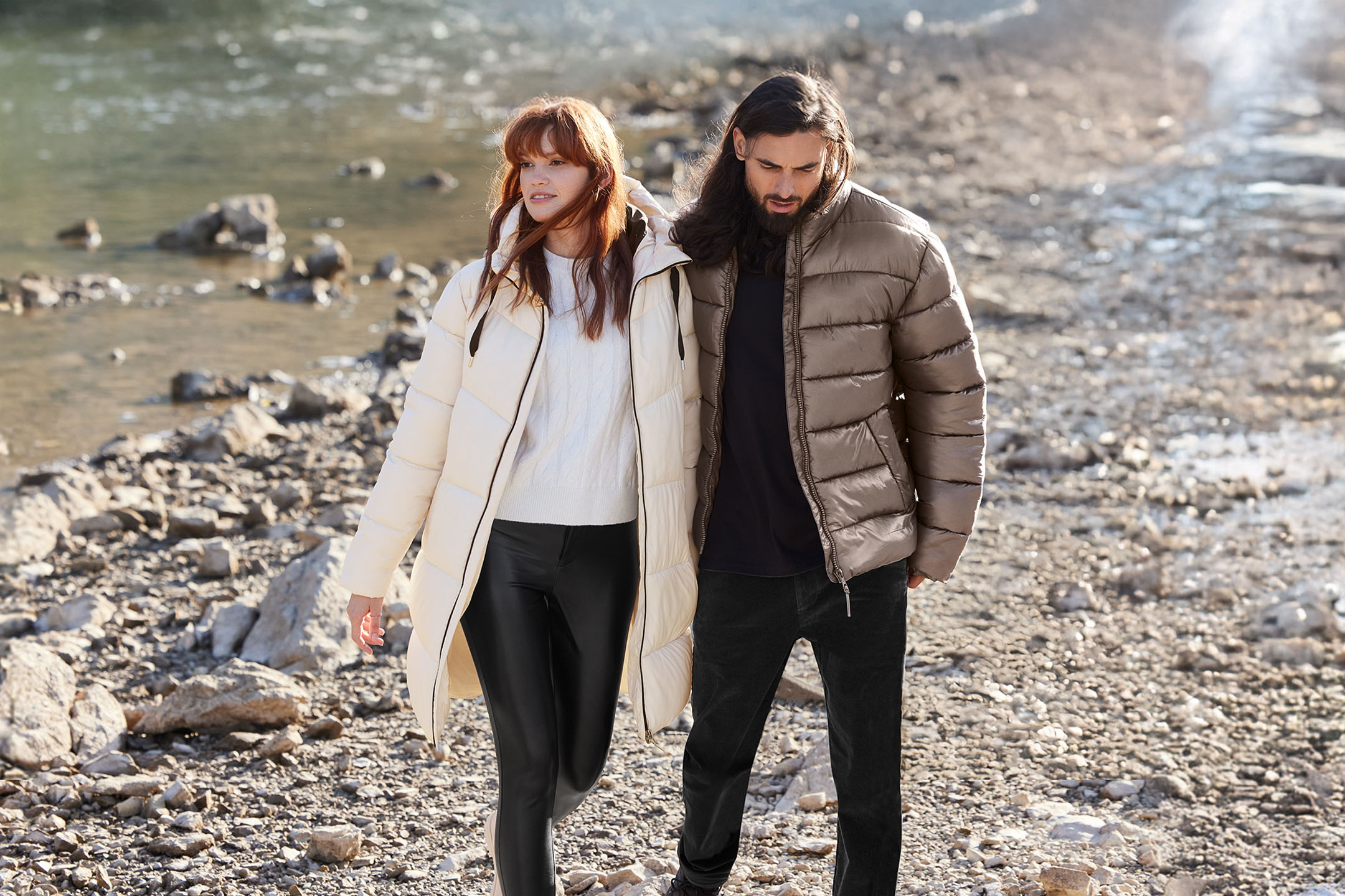 Couple in quilted jackets and dark trousers walking by the water.
