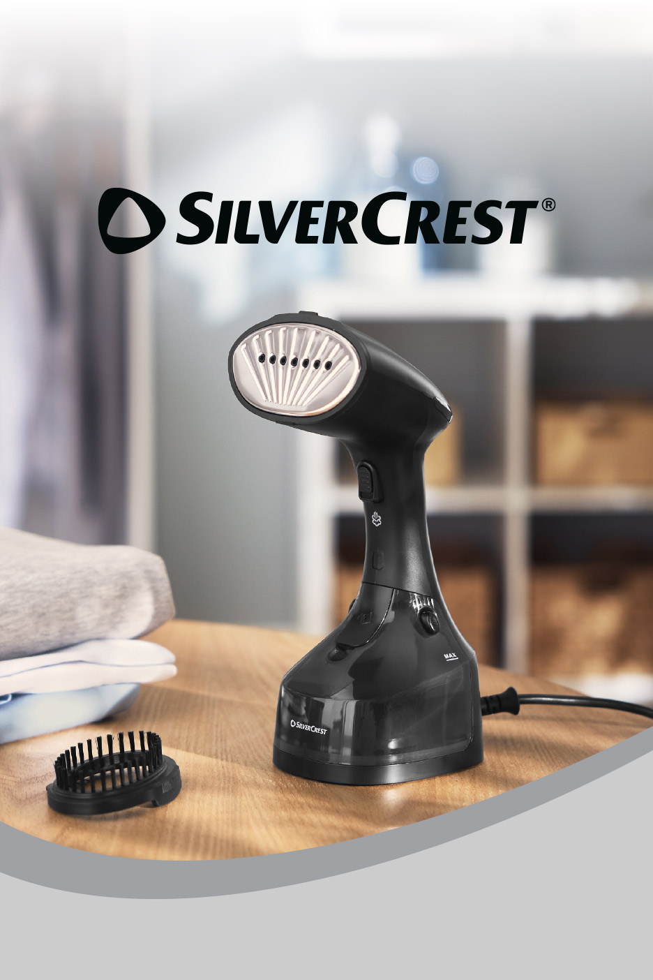 Black handheld garment steamer with a brush attachment on a wooden table next to folded clothes.