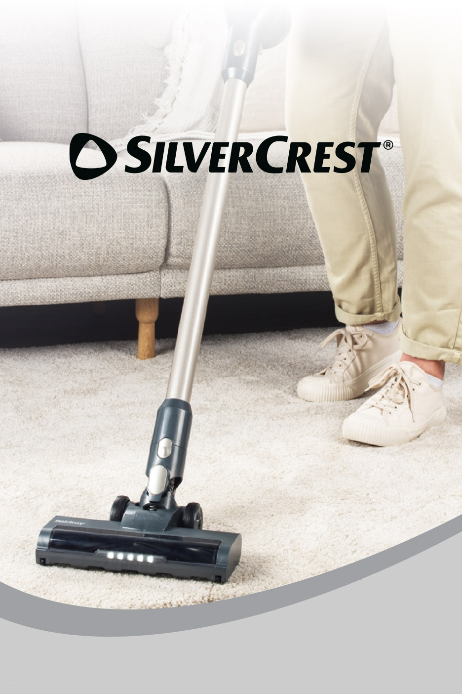 Cordless stick vacuum cleaner on a light carpet, with a person's legs and a sofa in the background.