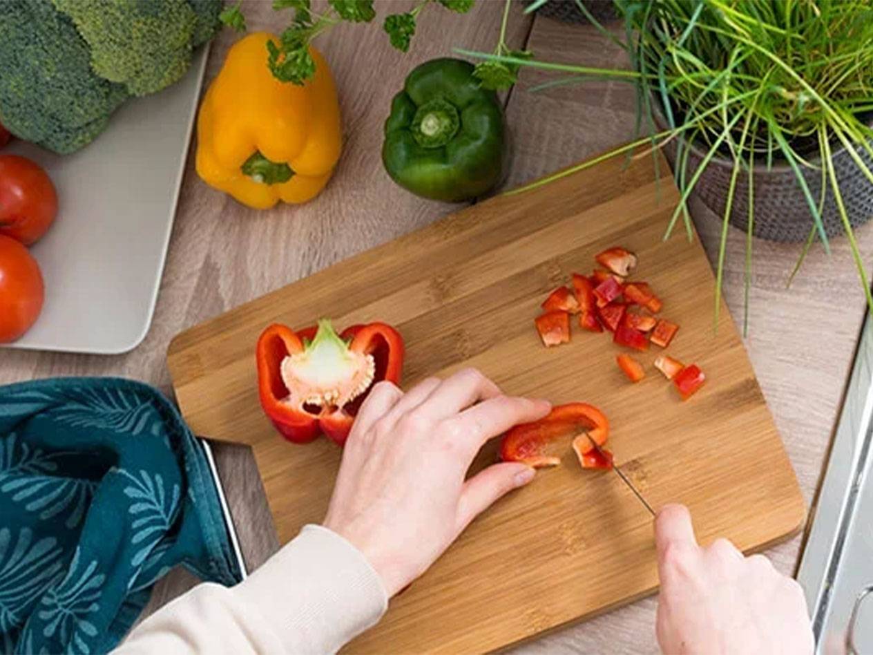 A person cuts red bell pepper on a wooden cutting board, with other vegetables around.