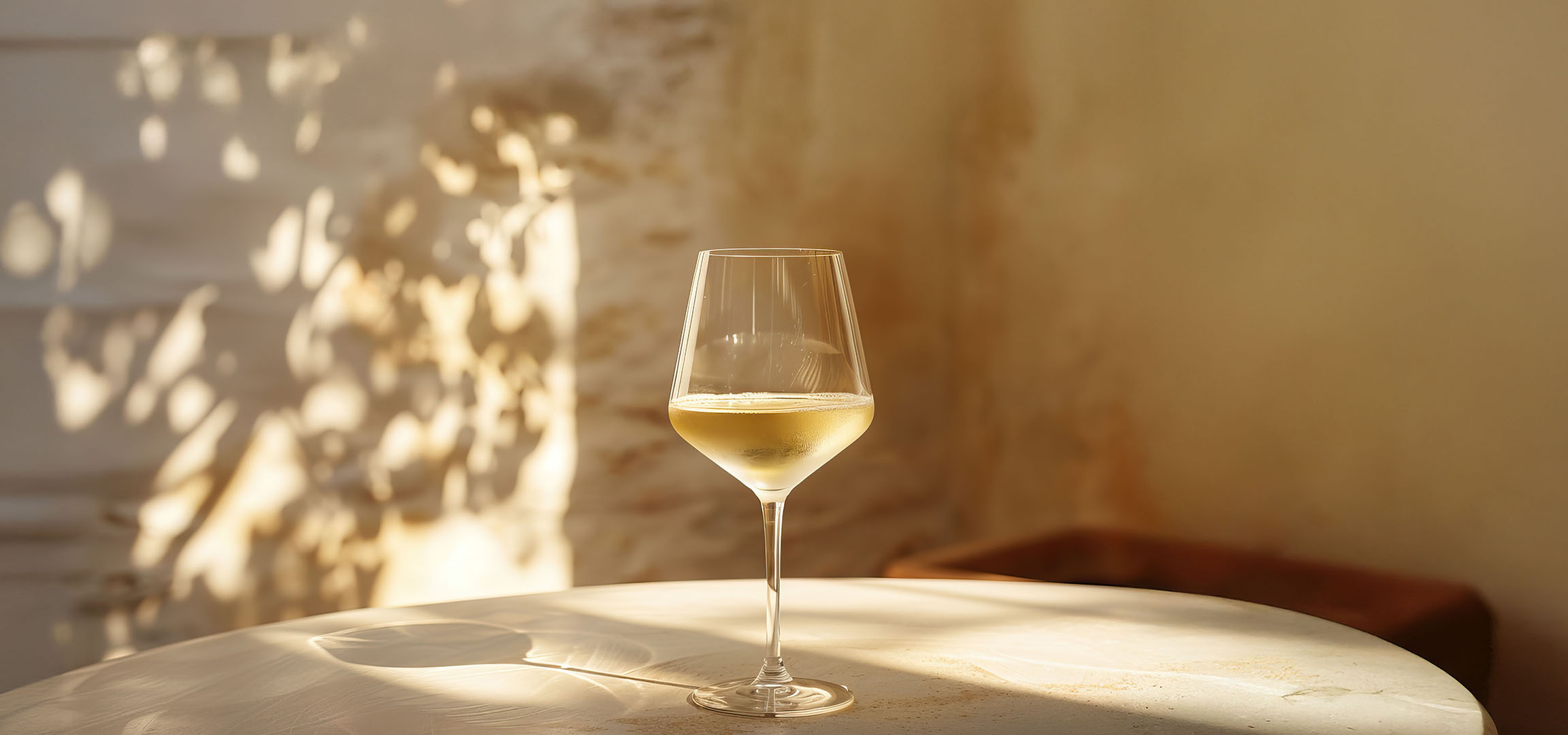 Glass of white wine on a table, with shadows of leaves and columns.