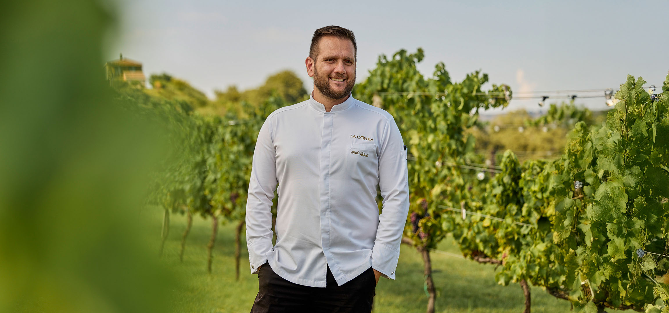 Smiling man in a white chef's jacket in a sunny vineyard.