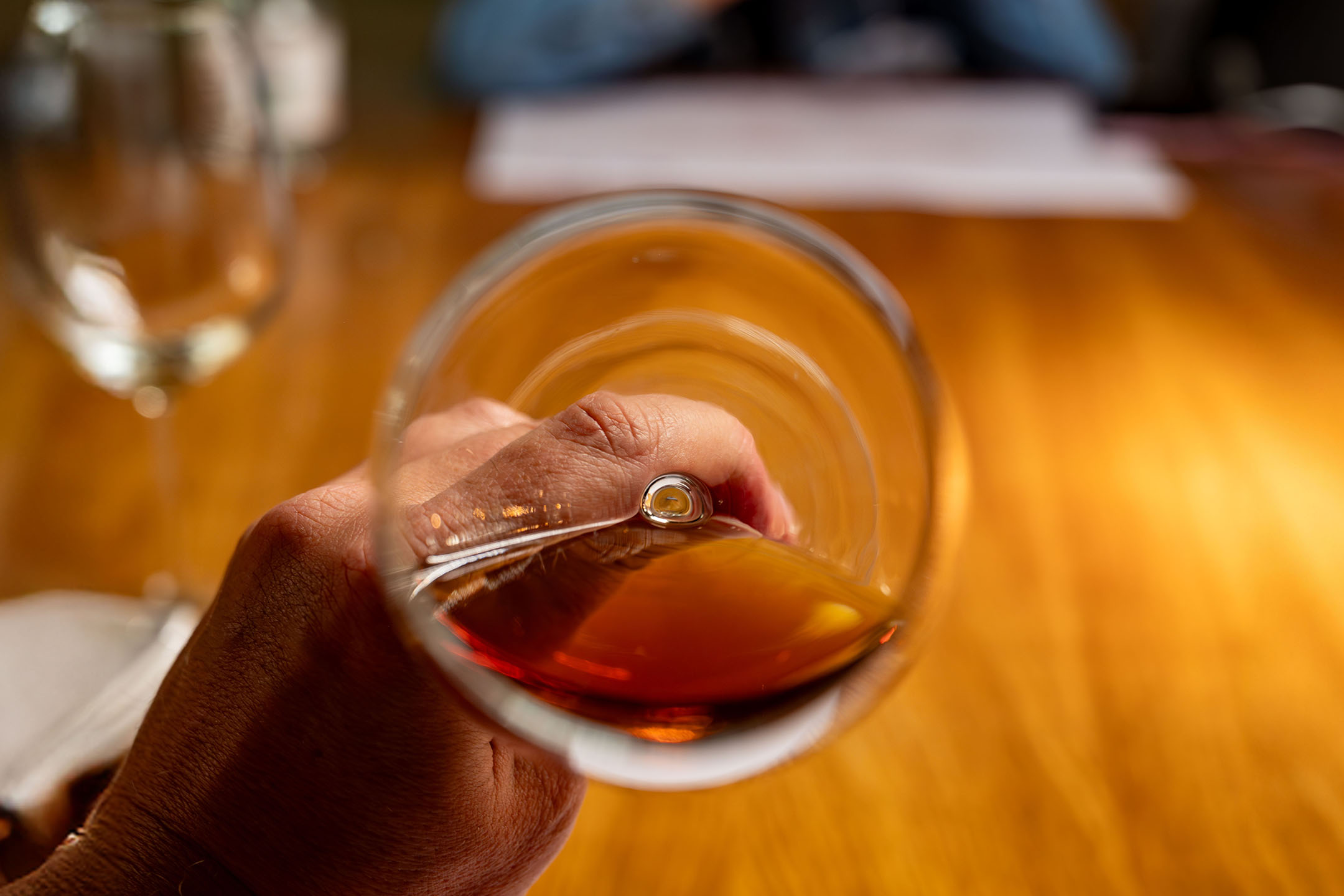 Hand holding a glass of amber liquid, possibly brandy or whisky, over a wooden table.