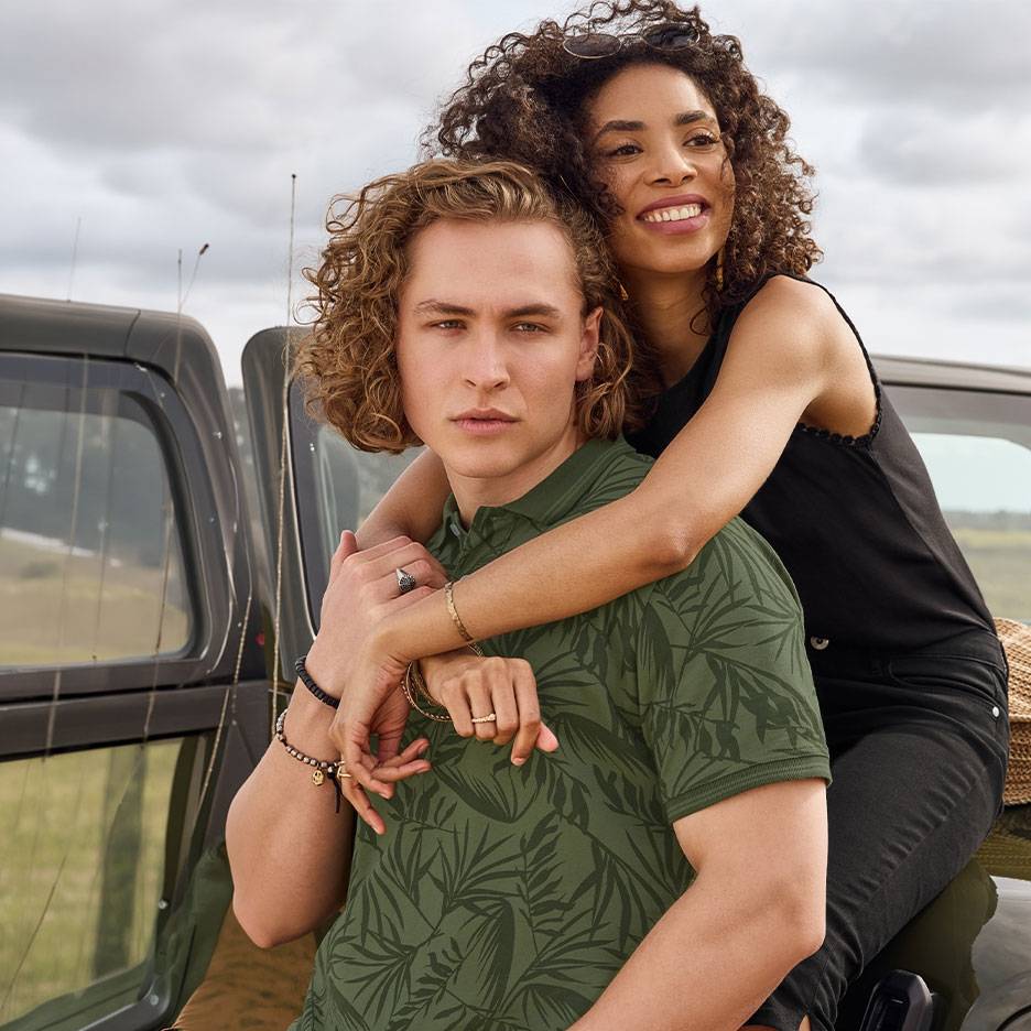 Smiling couple, he in a green leaf-patterned polo shirt, she in a black top, by a car.