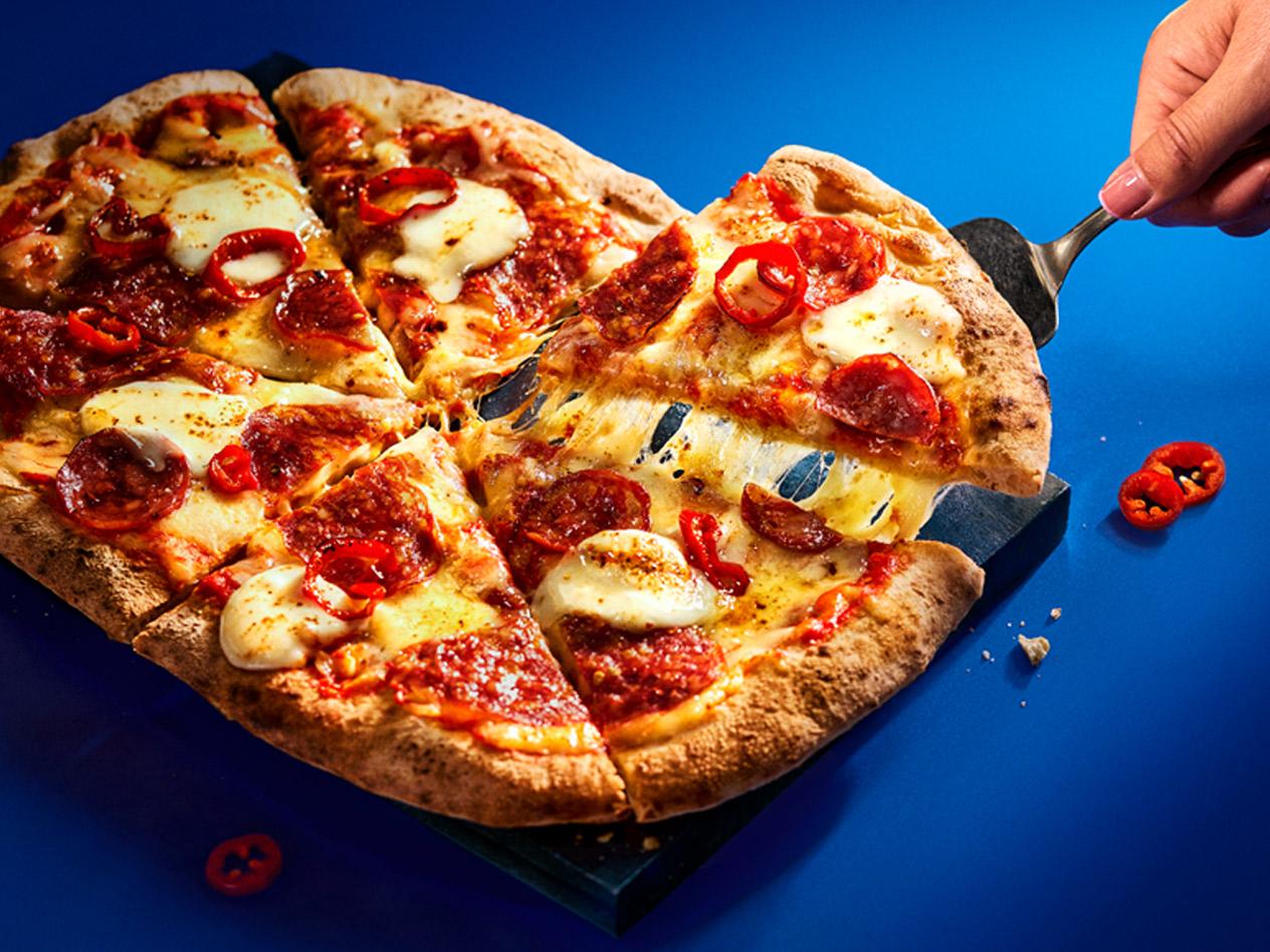 A hand lifting a slice of Deluxe hot pepperoni pizza with melting cheese on a blue background.