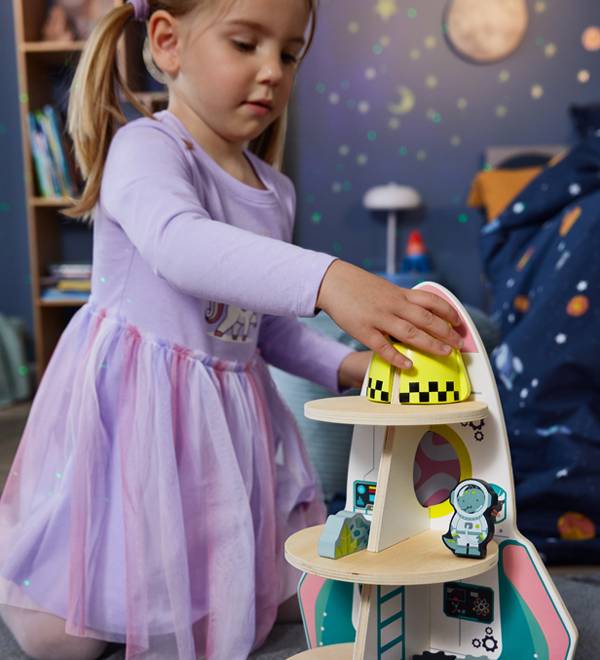 A young girl playing with a wooden rocket ship toy in a space-themed room.