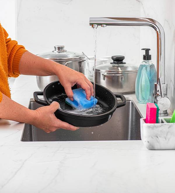 A person washing a black pan with a blue sponge under a kitchen faucet.