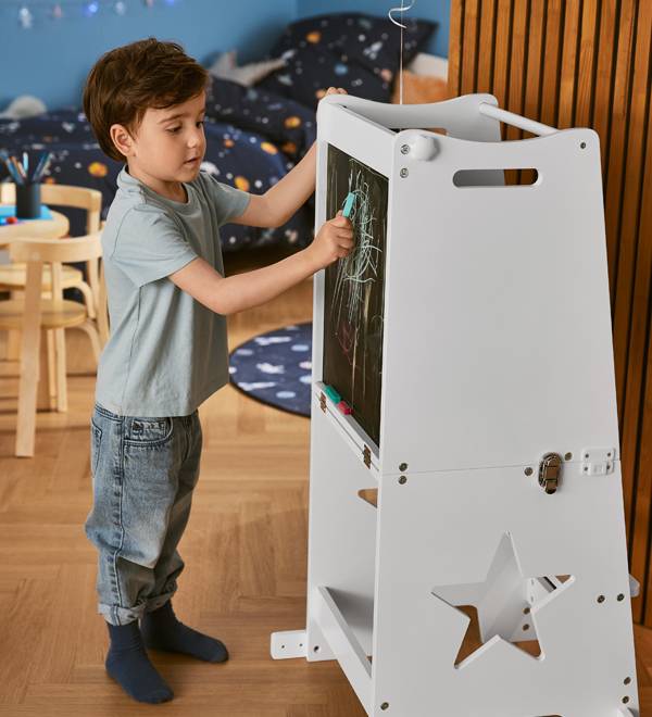 A young boy drawing on a white wooden learning tower with a chalkboard.