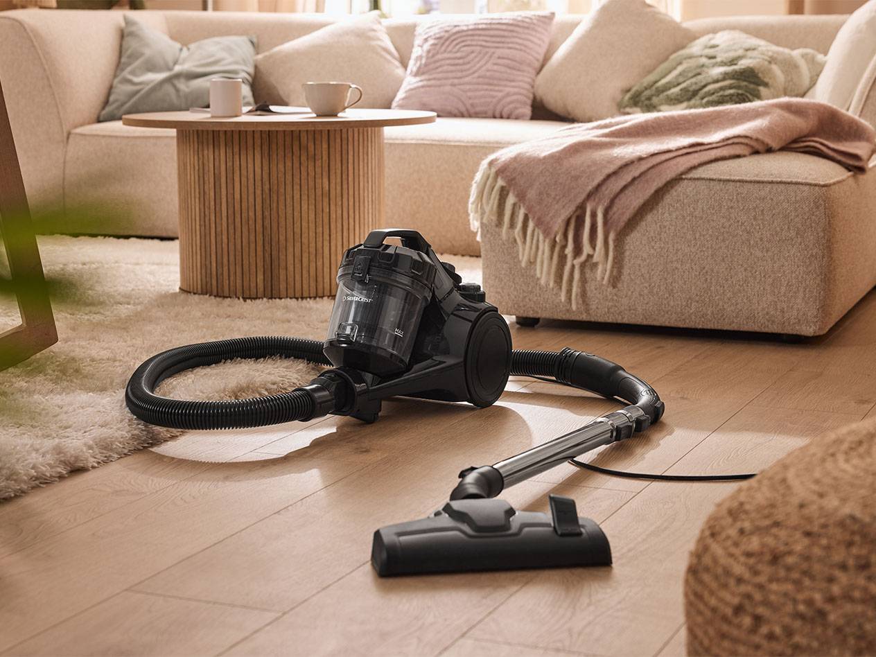 Black SilverCrest vacuum cleaner on wooden floor with a cozy sofa and coffee table in the background.