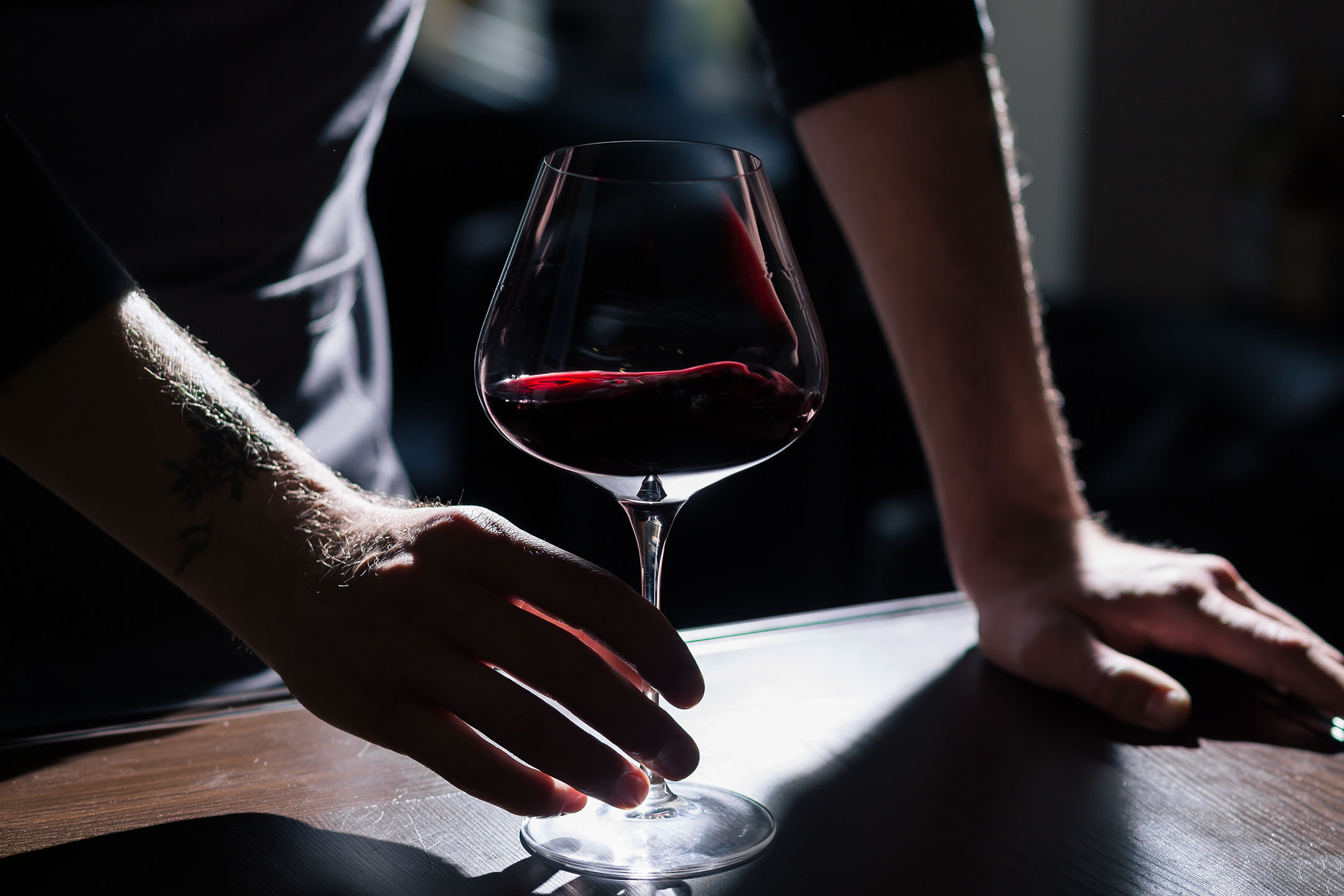 Tattooed hands of a man holding a glass of red wine on a wooden table.