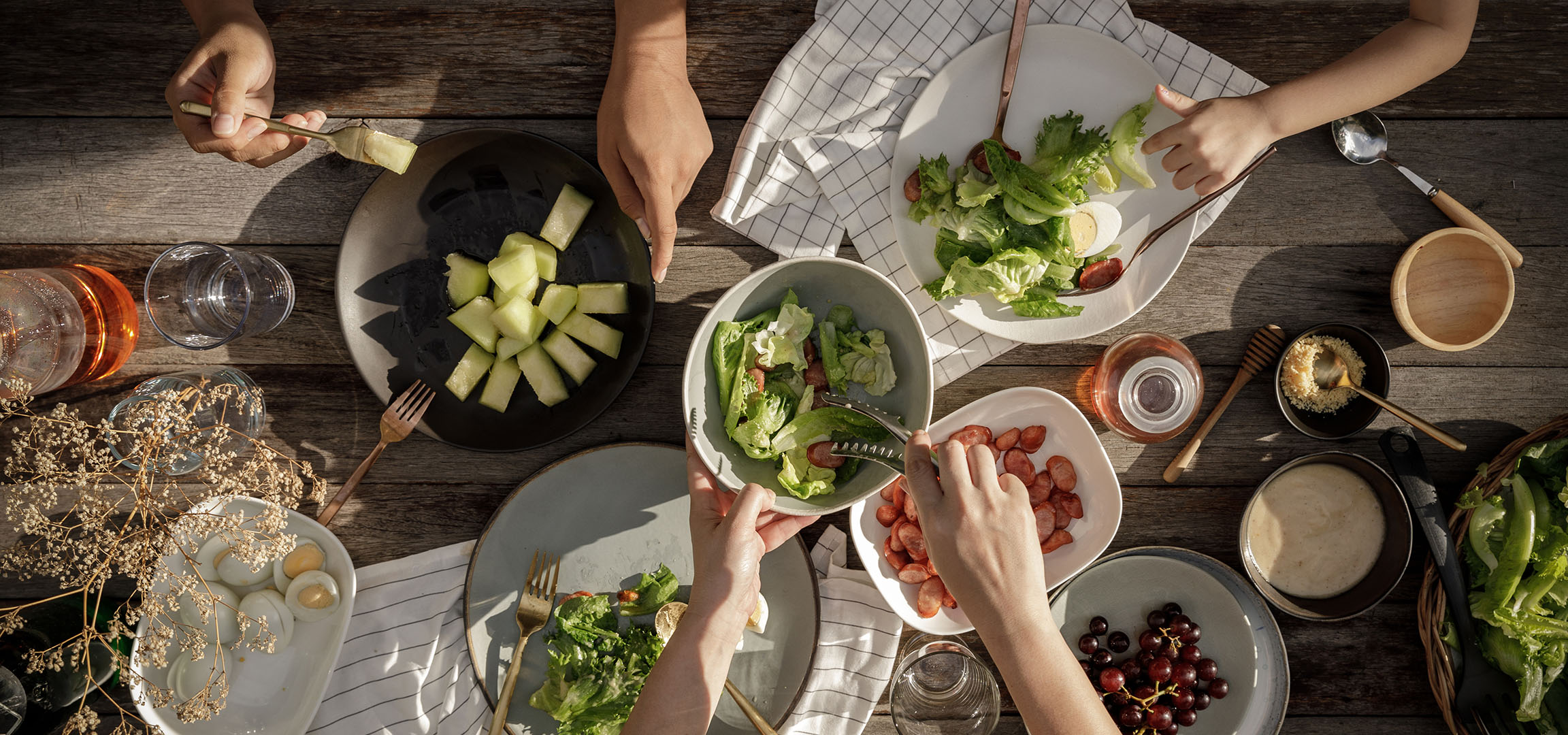 Overhead view of a table laden with salad, sausages, and melon, with people serving food.