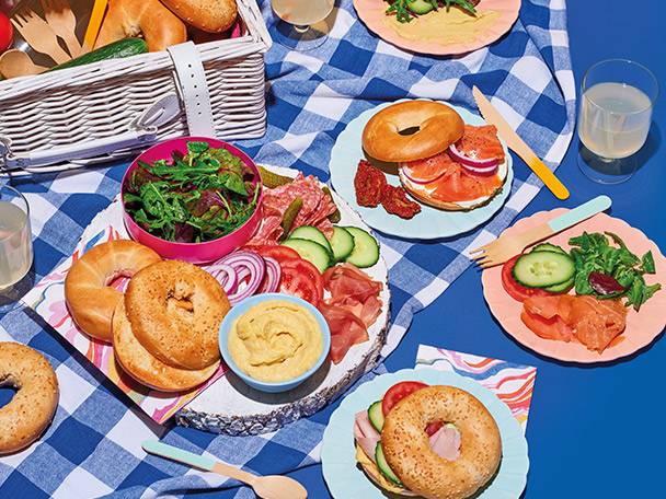 Picnic spread with bagels, smoked salmon, deli meats, salad, and hummus on a blue checkered blanket.