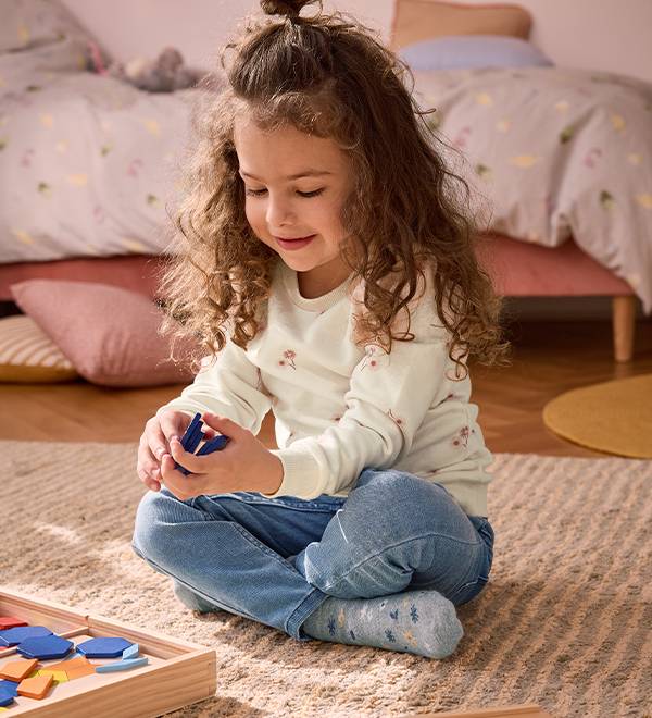 A young girl in a long-sleeved shirt and jeans playing with a wooden puzzle.
