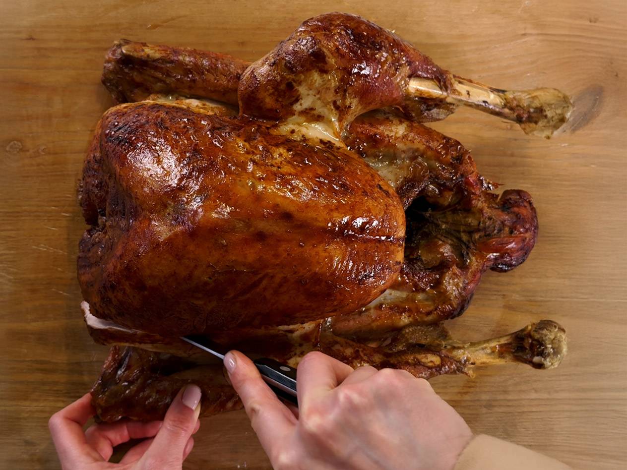 Hands carving a roasted turkey on a wooden cutting board.