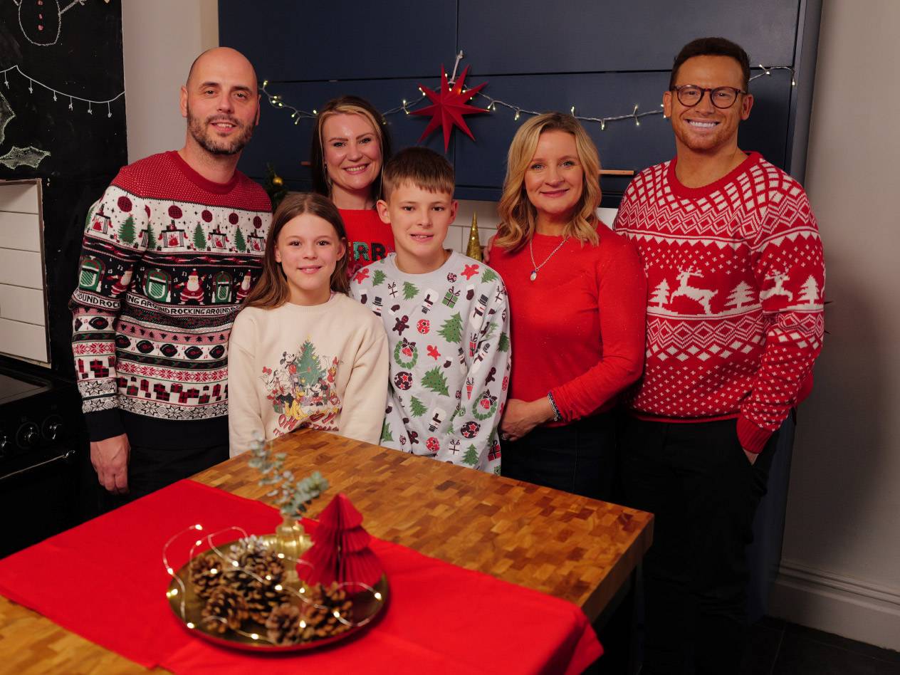 Family wearing Christmas jumpers, standing around a table with festive decorations.