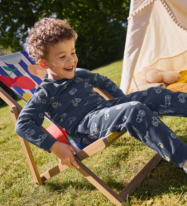 A young boy in a blue tractor-patterned pajama set sits in a colorful striped deck chair.