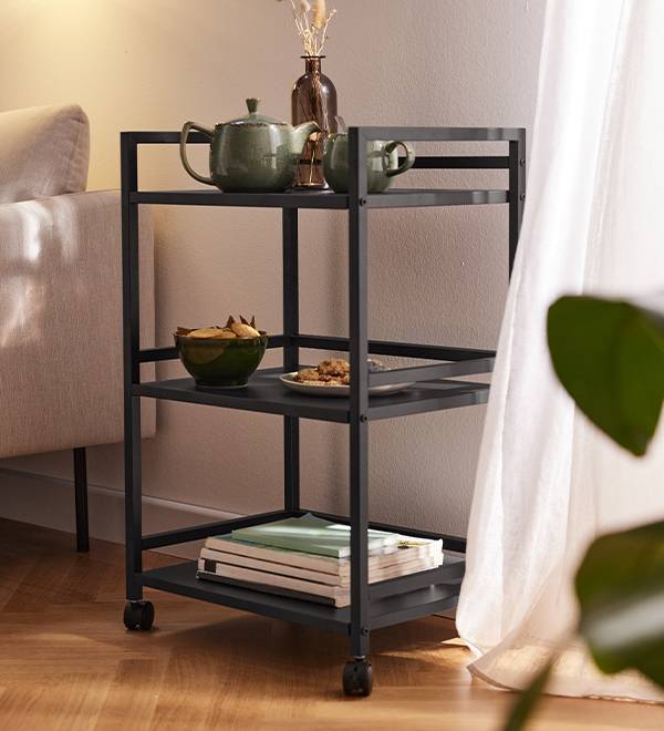 A black metal rolling cart with three shelves, holding a teapot, snacks, and books.