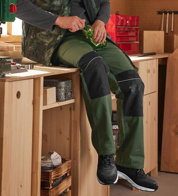 Man in workwear sitting on a workbench, holding a green bottle.