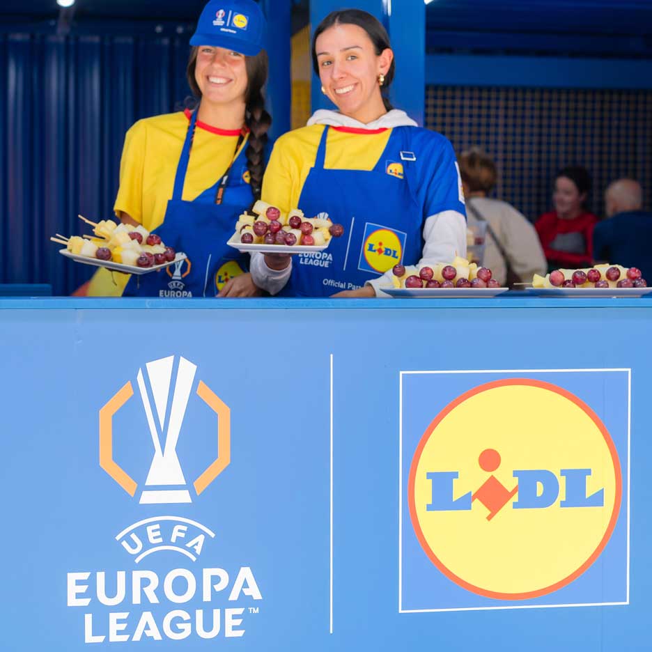 Two smiling women in Lidl aprons offer fruit on skewers at a UEFA Europa League event.