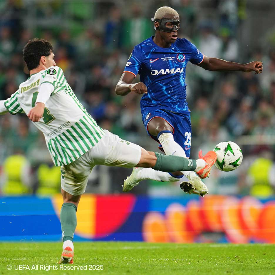 Two soccer players battle for the ball on the field, one in white and green, the other in blue with a mask.