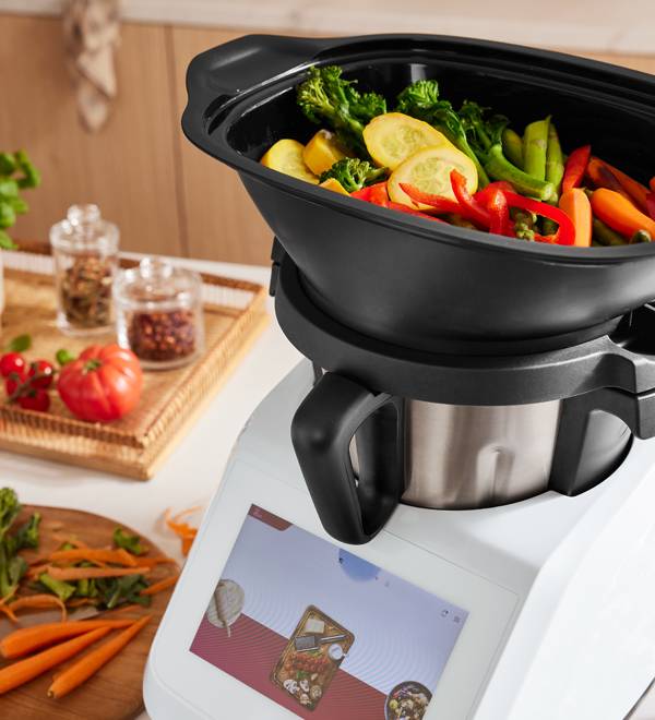 A food processor with a steaming basket full of colorful vegetables, with ingredients nearby.